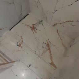 Andrii Kulchenko - Wykończona podłoga i ściany w łazience wyłożone białymi płytkami imitującymi marmur z rdzawymi żyłkami, widoczny odpływ liniowy i fragment obuwia osoby robiącej zdjęcie.