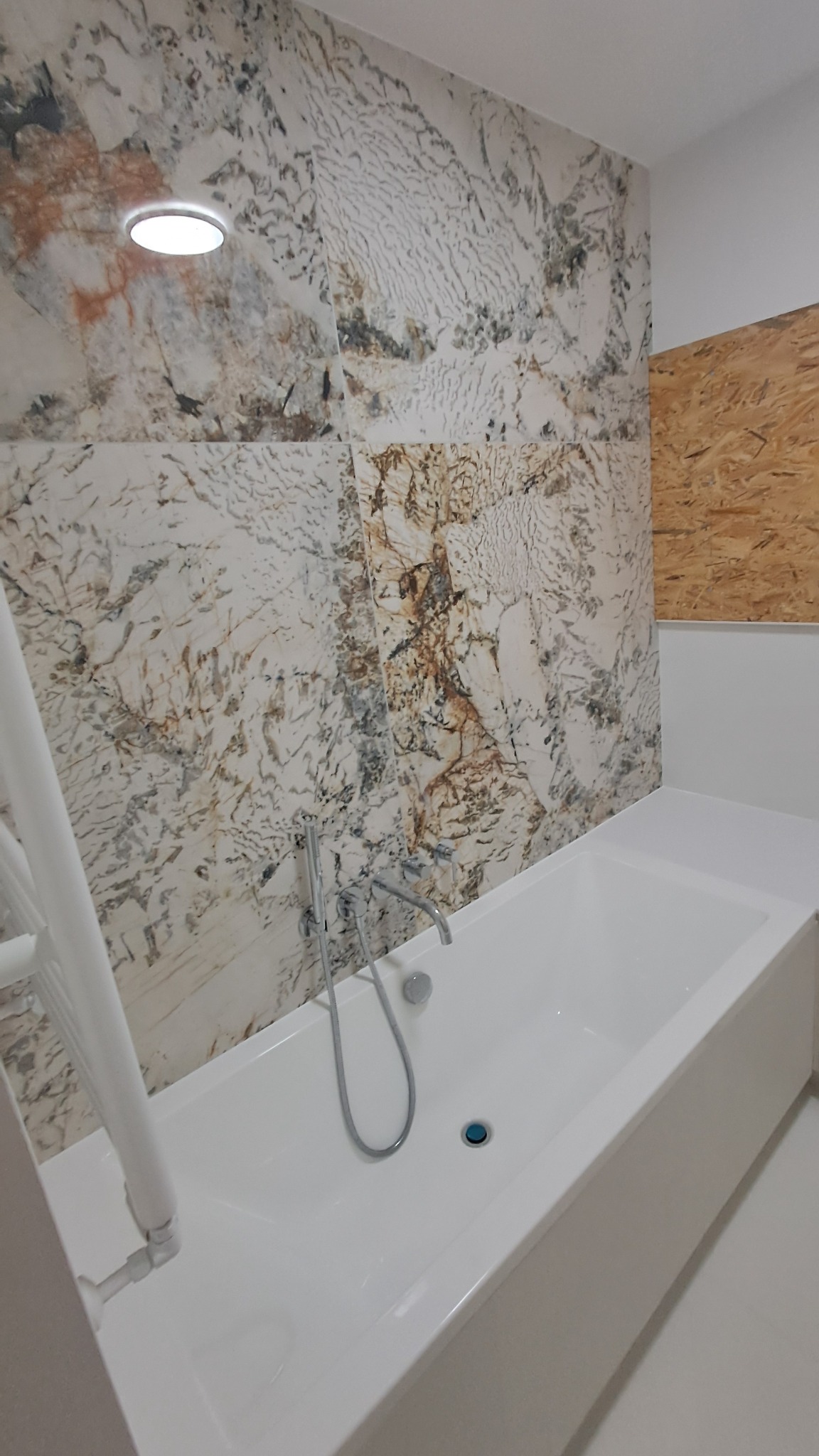 Nowoczesna łazienka z wanną obłożoną białymi płytkami, ściana wykończona płytkami imitującymi marmur z brązowymi żyłami, widoczny grzejnik drabinkowy i fragment ściany z płyt OSB.
