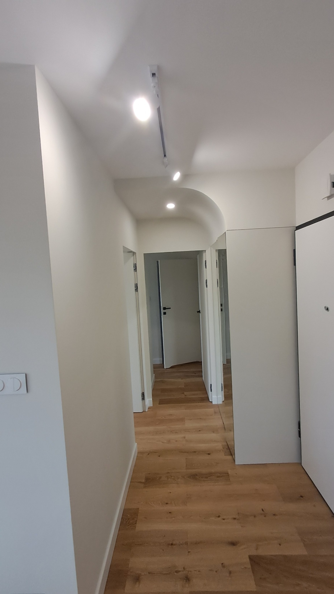 Nowoczesny korytarz z drewnianą podłogą, białymi ścianami i sufitowym oświetleniem szynowym, widok na otwarte drzwi do innych pomieszczeń.