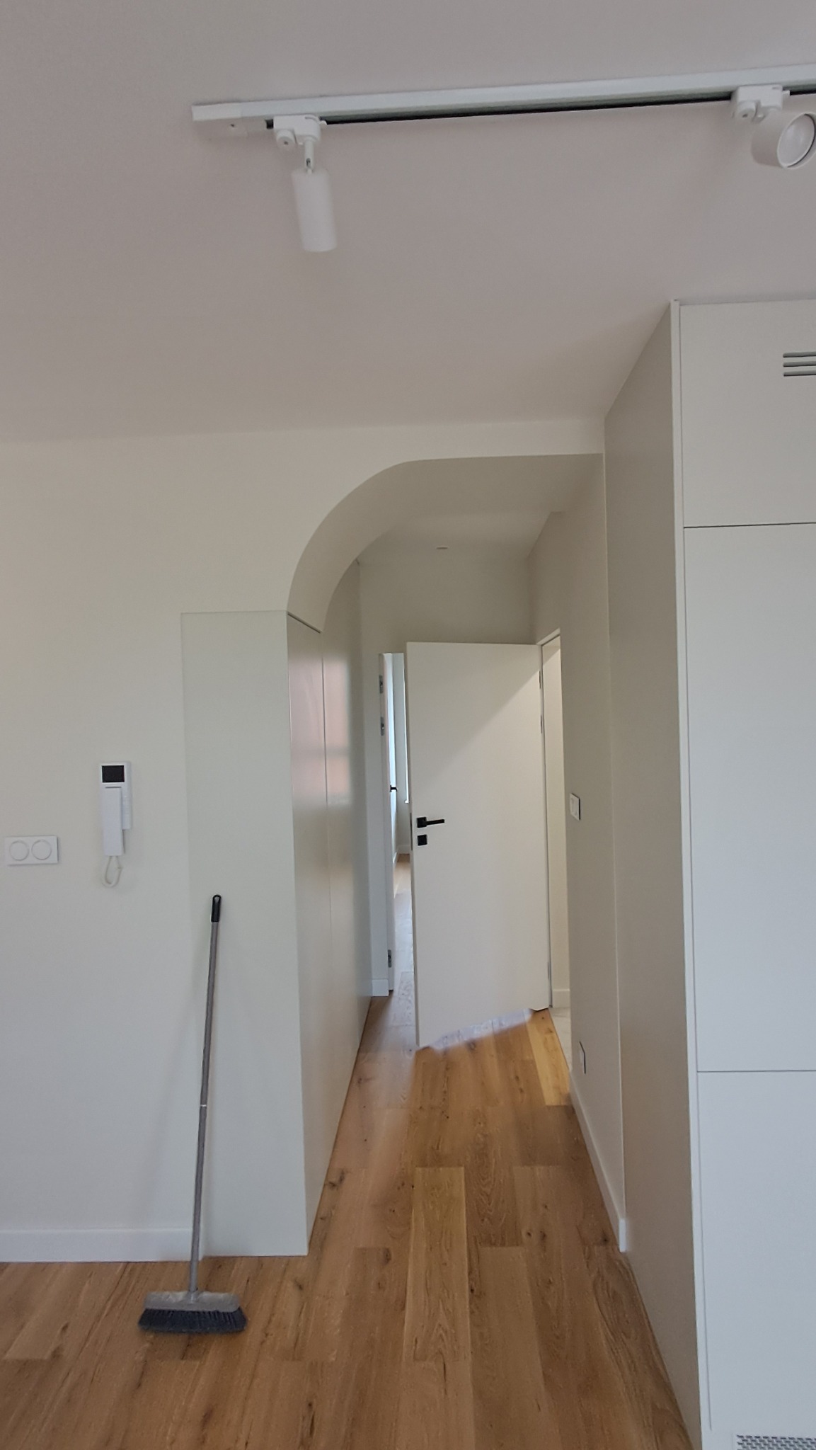 Wnętrze mieszkania po remoncie: świeżo pomalowane białe ściany, drewniana podłoga, widoczna miotła przy ścianie i oświetlenie punktowe na suficie.