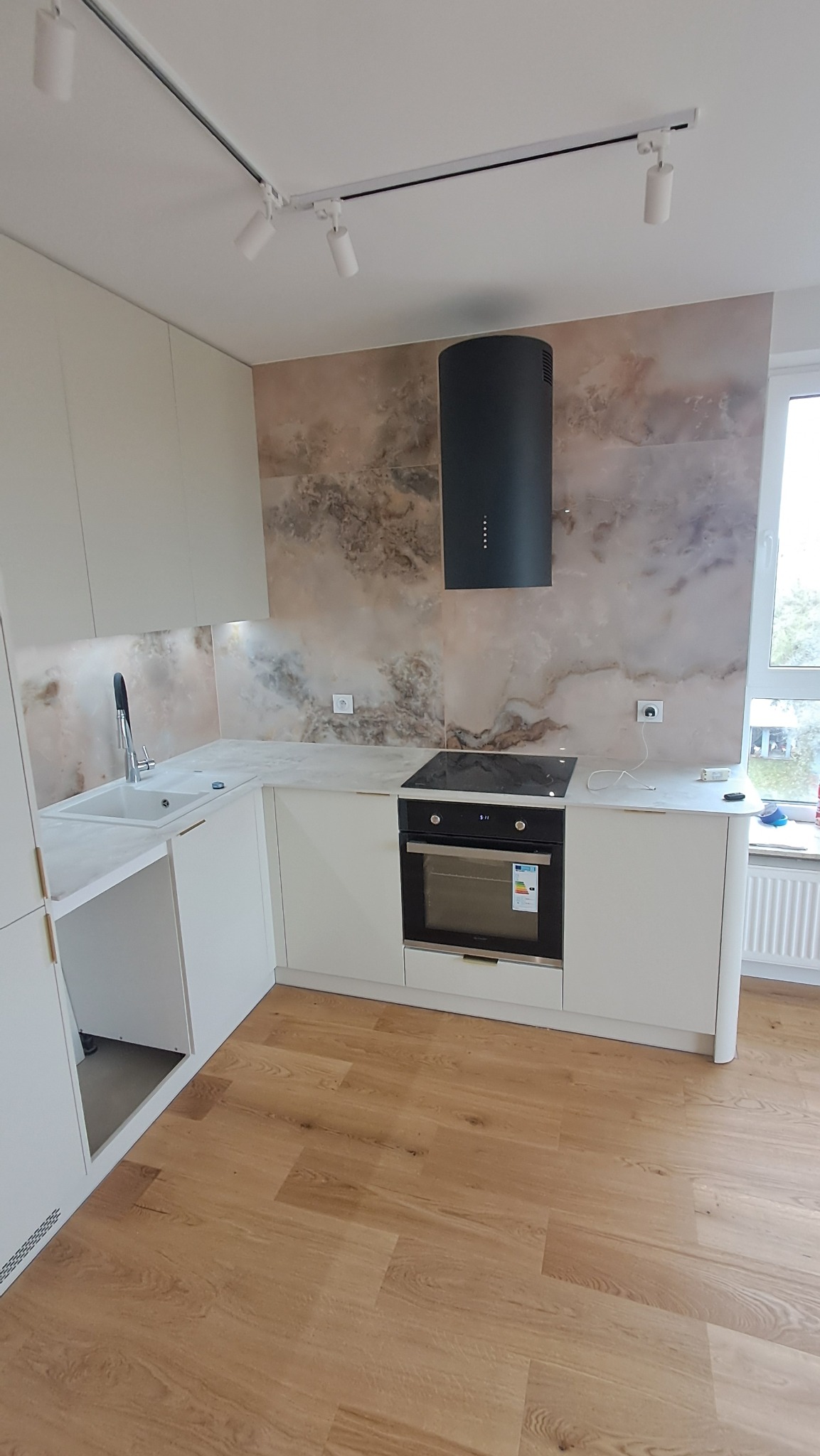 Nowoczesna kuchnia z białymi szafkami, marmurowym blatem i okładziną ścienną o wzorze marmuru, czarnym okapem cylindrycznym oraz drewnianą podłogą z paneli.