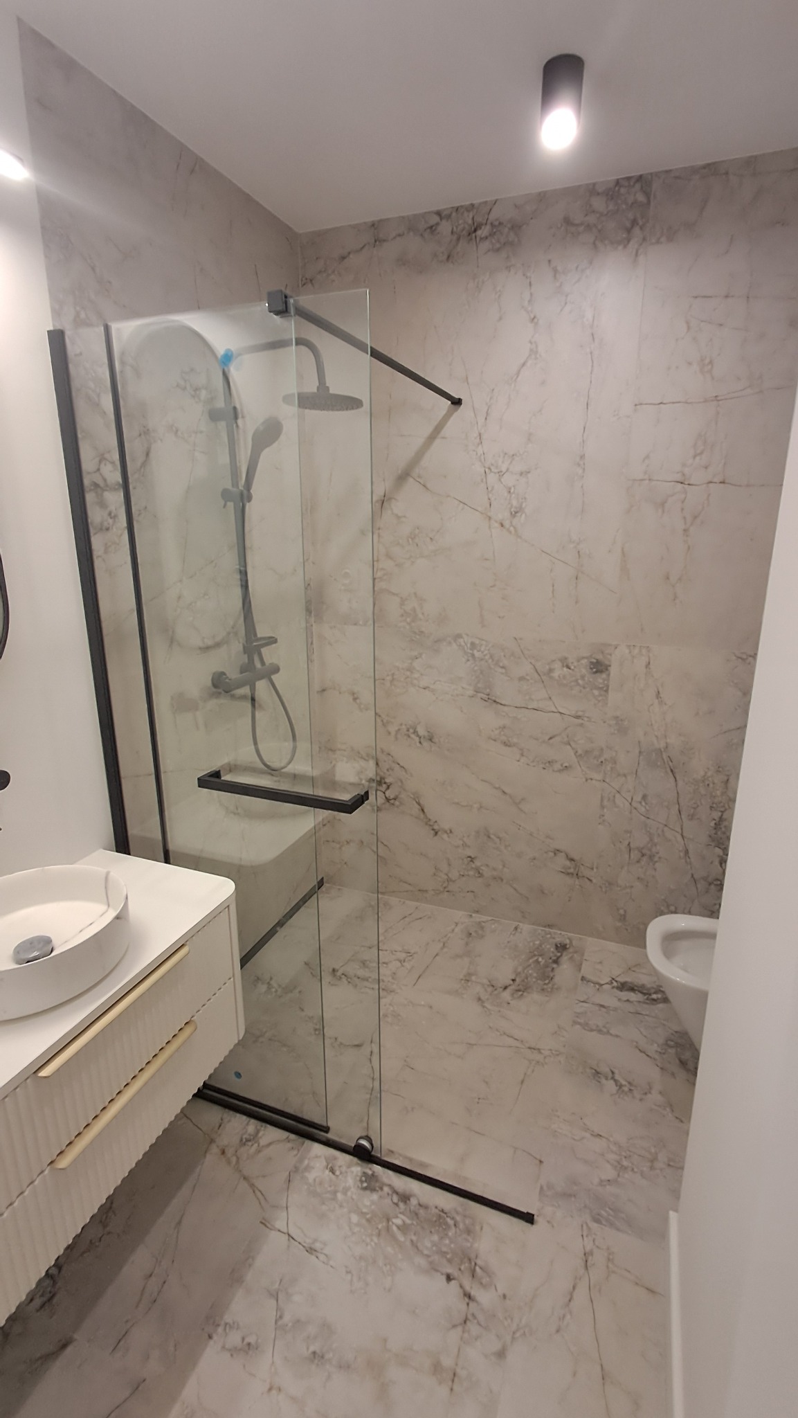 Nowoczesna łazienka z kabiną prysznicową typu walk-in z czarnymi profilami, umywalką nablatową na białej szafce z frezowanymi frontami i marmurowymi płytkami.