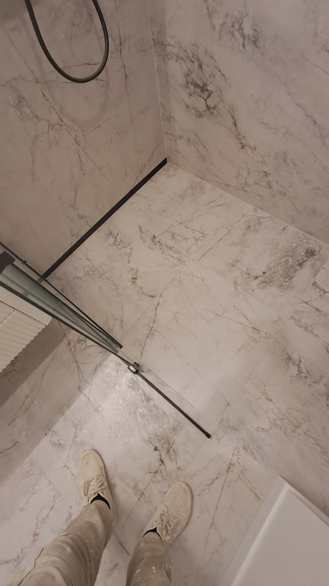 Wykończona kabina prysznicowa z marmurowymi płytkami, widok z góry na podłogę i fragment osoby w ubrudzonych farbą spodniach i butach.