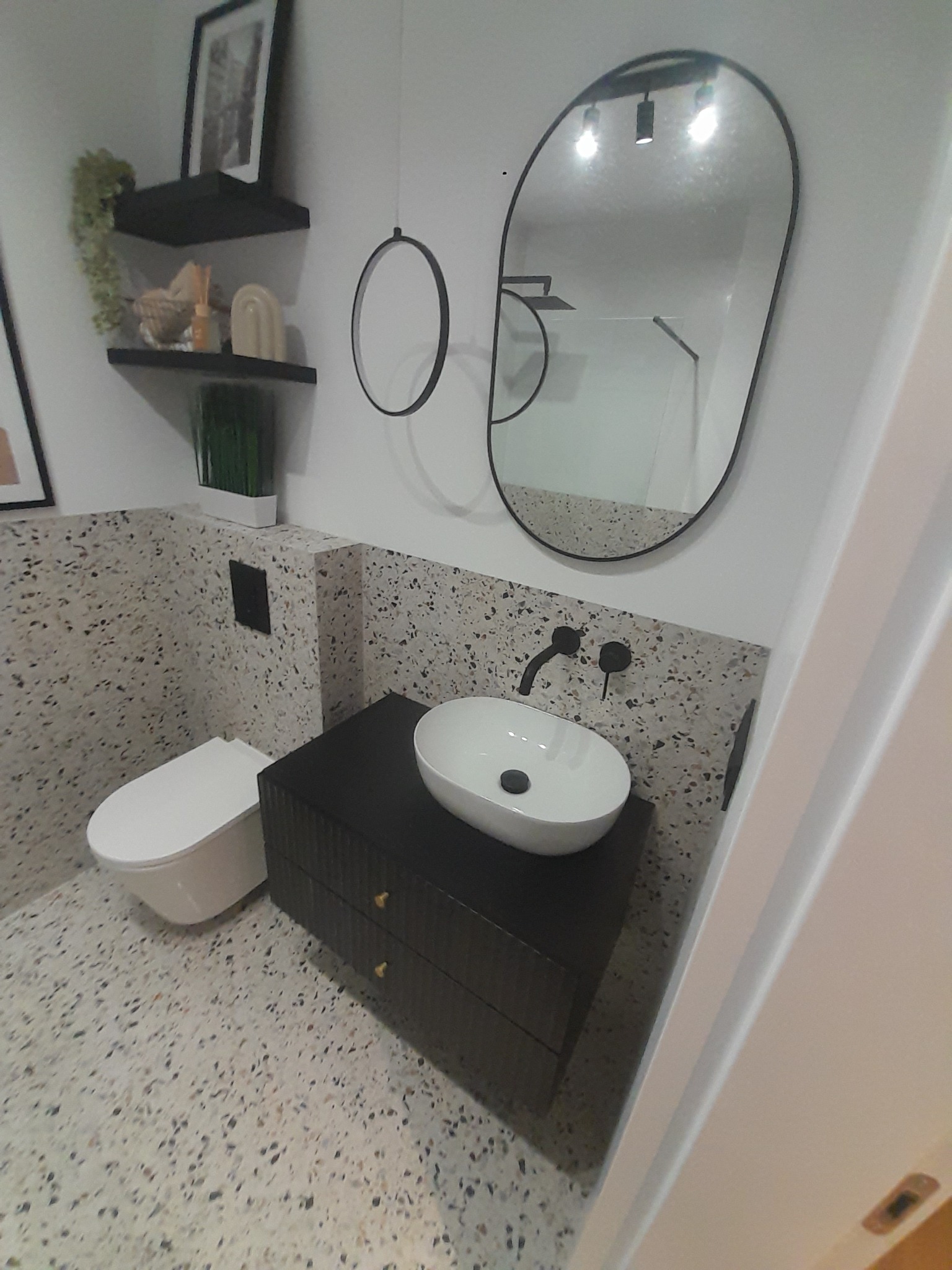 Nowoczesna łazienka z umywalką nablatową, owalnym lustrem z czarną ramą, toaletą wiszącą i płytkami lastryko na ścianach i podłodze.