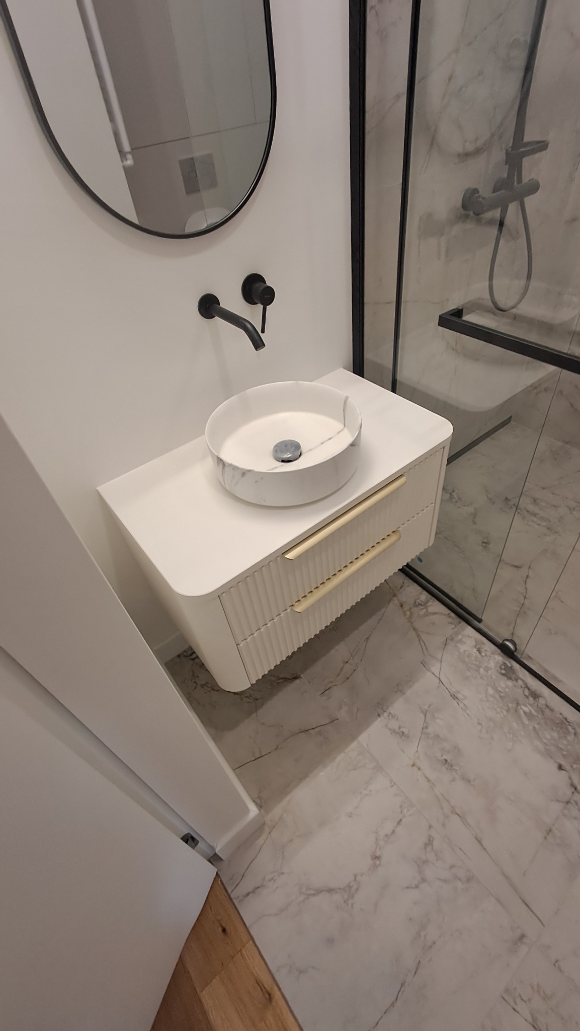 Nowoczesna łazienka z okrągłą umywalką marmurkową na białej szafce z ryflowanymi frontami i złotymi uchwytami, czarna armatura i owalne lustro, kabina prysznicowa z czarnymi ramami, marmurowe...