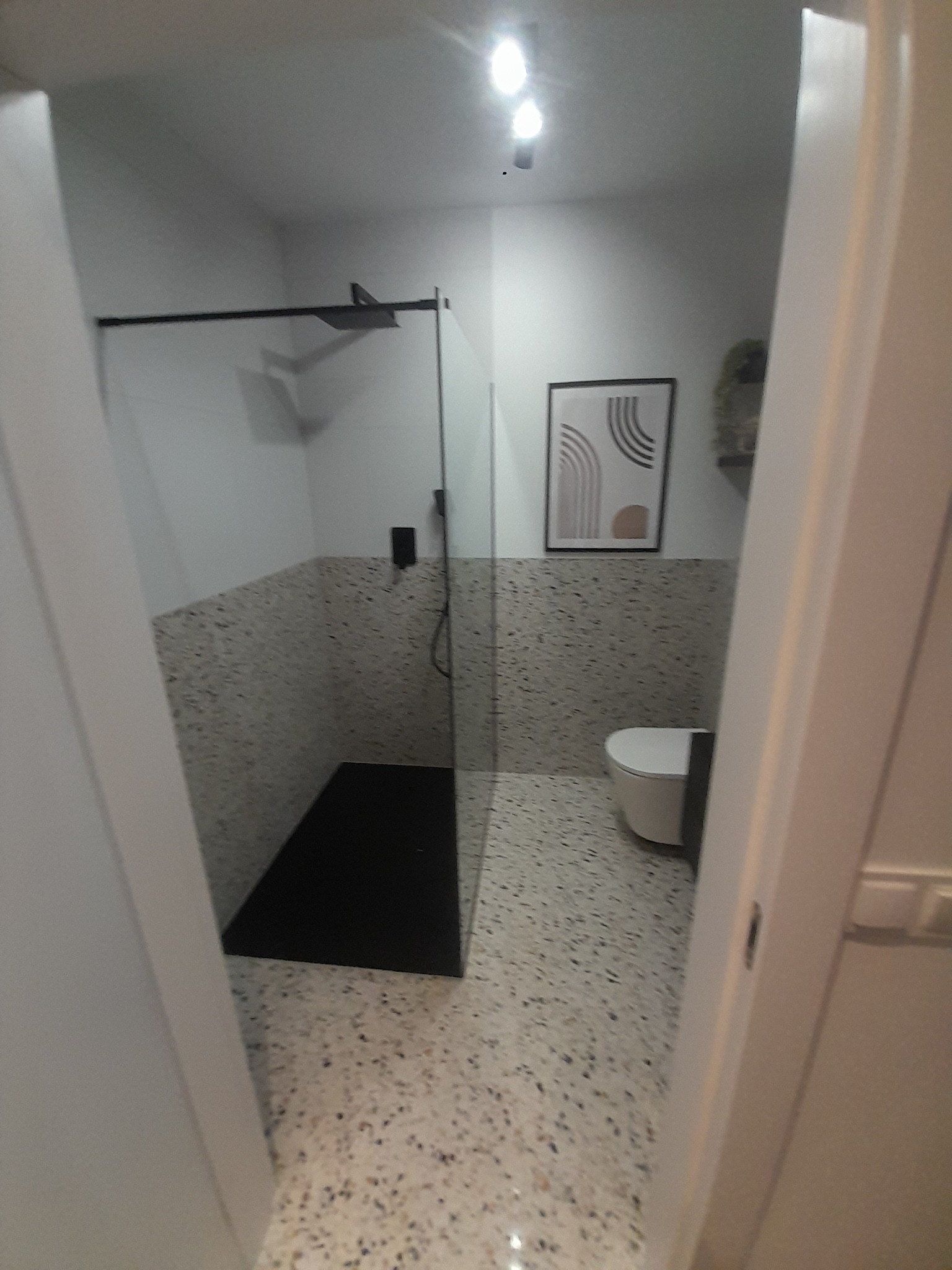 Nowoczesna łazienka z kabiną prysznicową typu walk-in, wykończona płytkami lastryko w odcieniach szarości i bieli, czarną armaturą i minimalistycznym plakatem na ścianie.