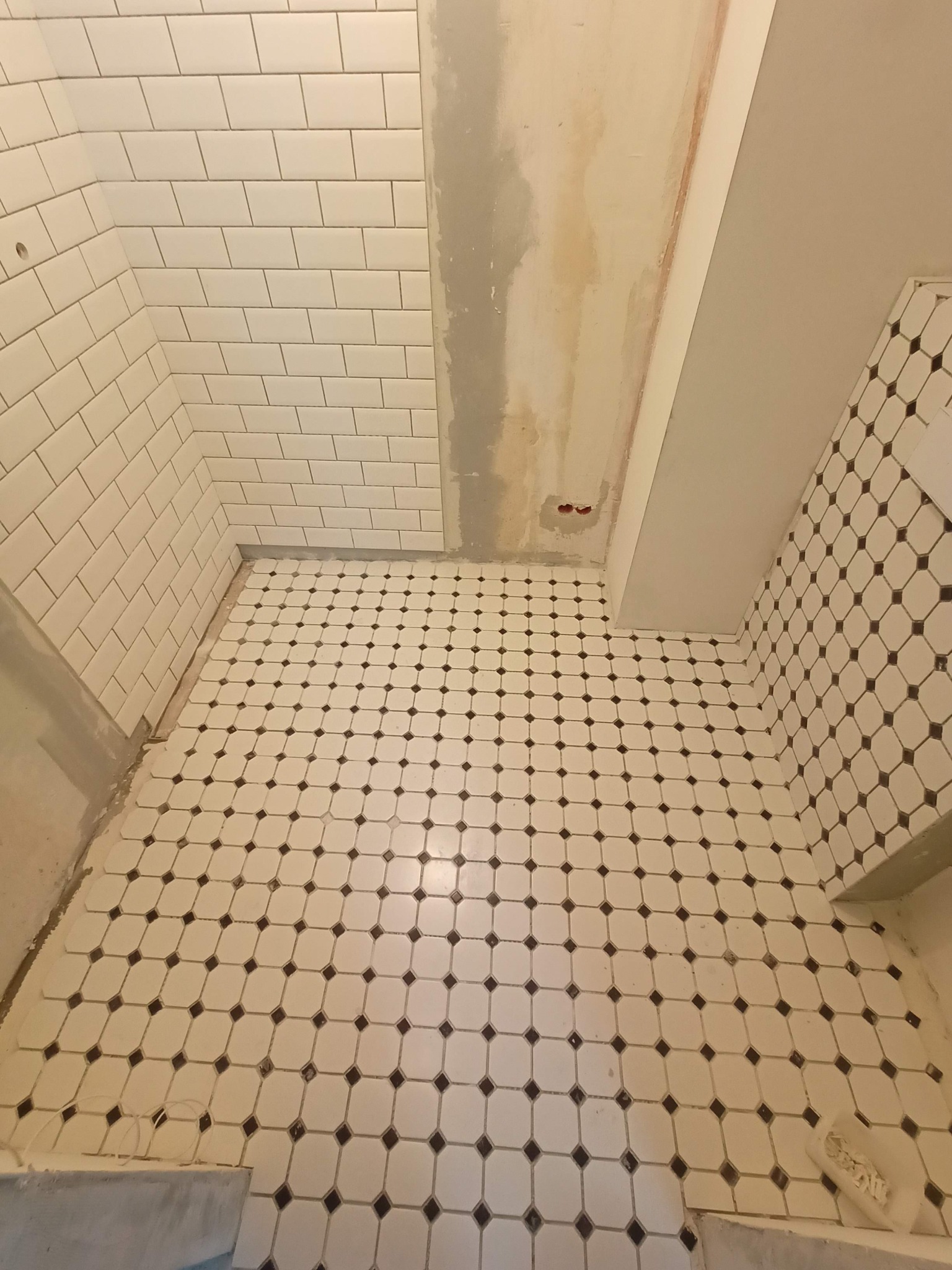 Łazienka w trakcie remontu: białe, prostokątne płytki na ścianach ułożone w cegiełkę, podłoga wyłożona biało-czarną mozaiką, ściana częściowo przygotowana do położenia płytek, widoczne puszki...