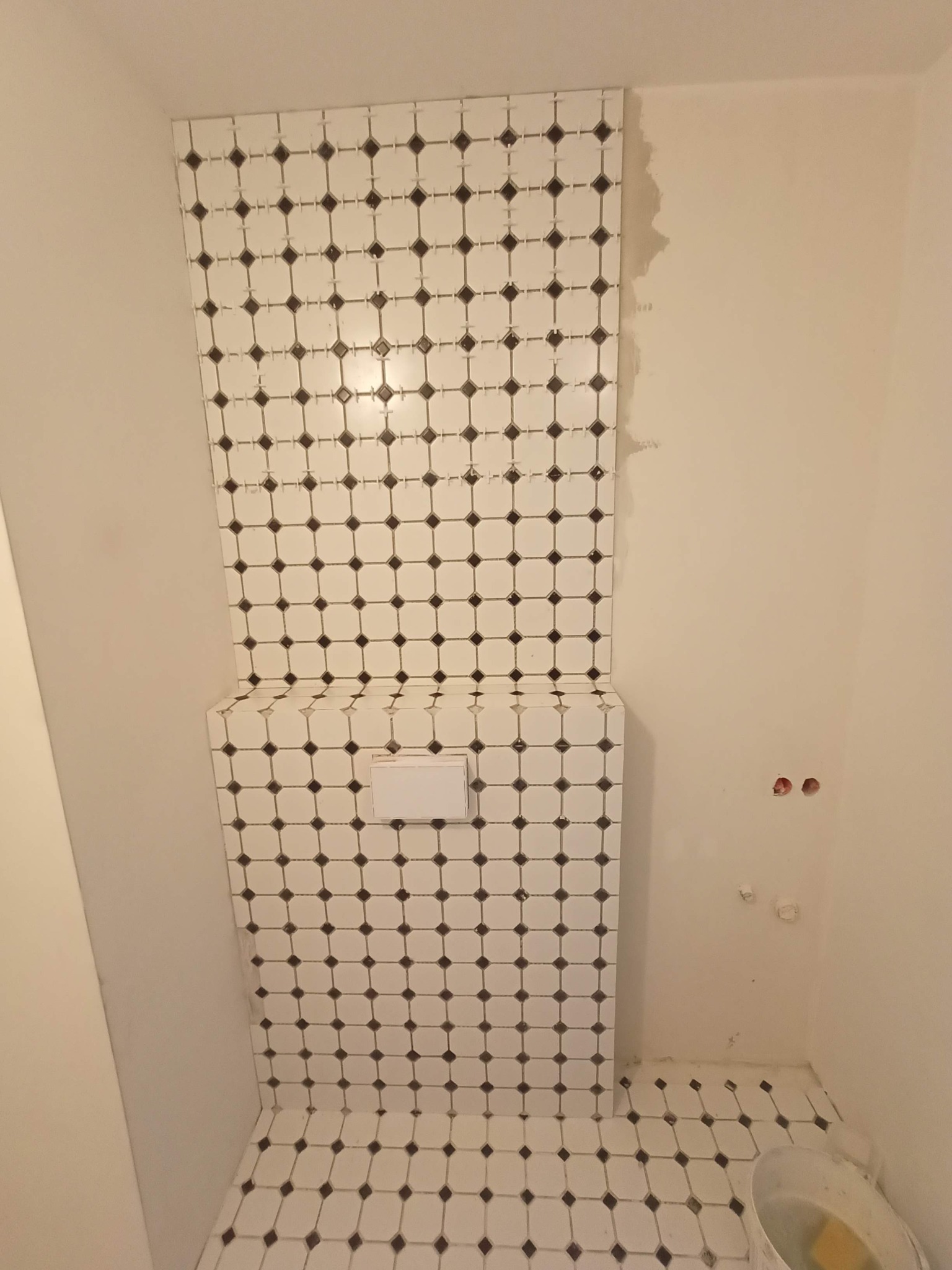 Montaż mozaiki z białych płytek z czarnymi akcentami wokół podwieszanego sedesu w trakcie remontu łazienki, widoczne krzyżyki dystansowe i pozostałości kleju.