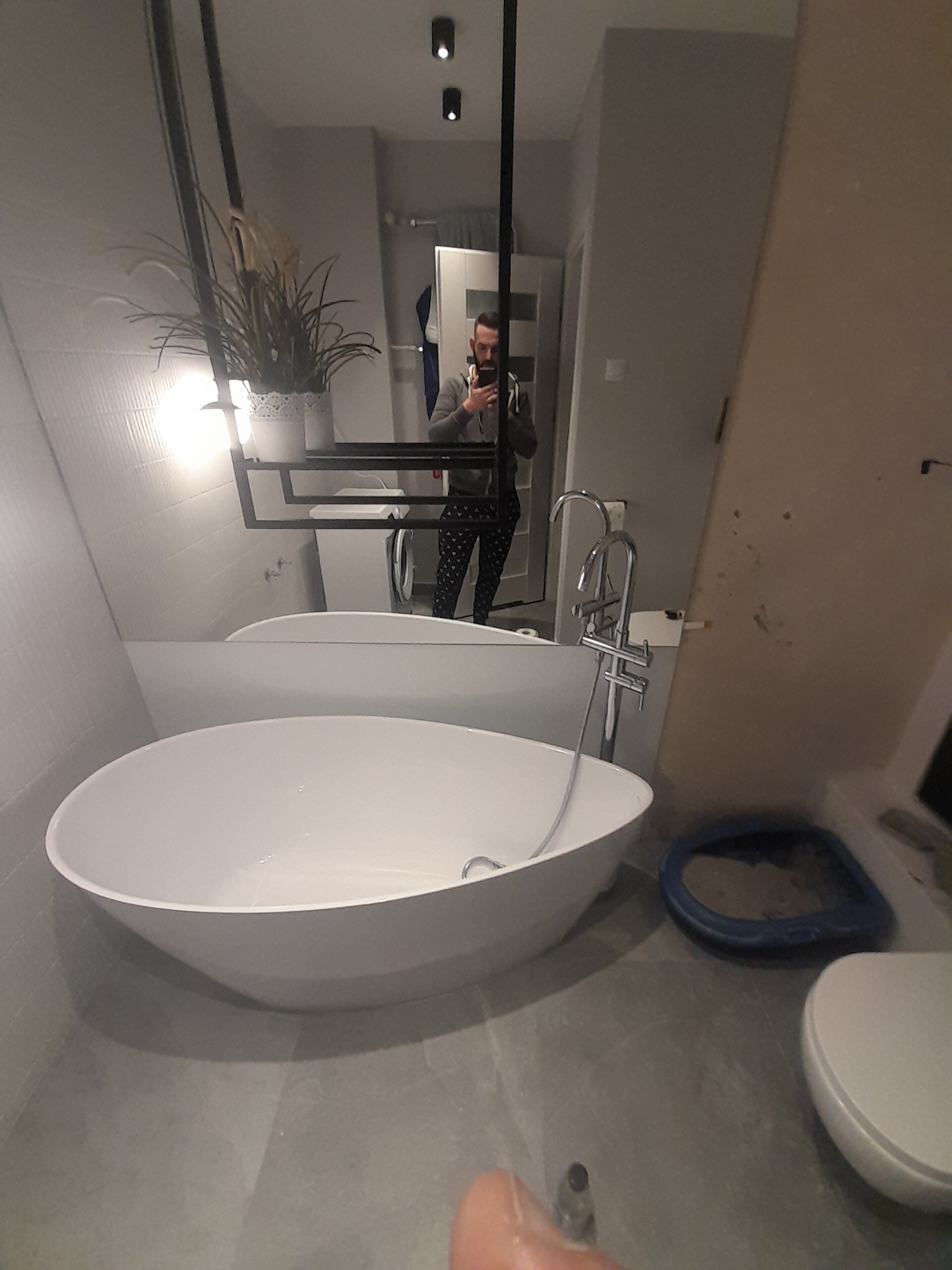 Nowoczesna łazienka z wolnostojącą białą wanną, szarą glazurą, toaletą i odbiciem osoby robiącej zdjęcie w lustrze.