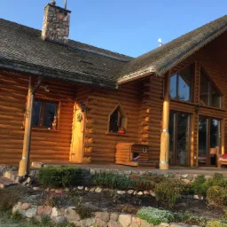 Wykończony dom z bali p&oacute;łokrągłych z dachem krytym gontem drewnianym, widoczny kamienny komin i elementy dekoracyjne dachu, ogr&oacute;dek przed domem.