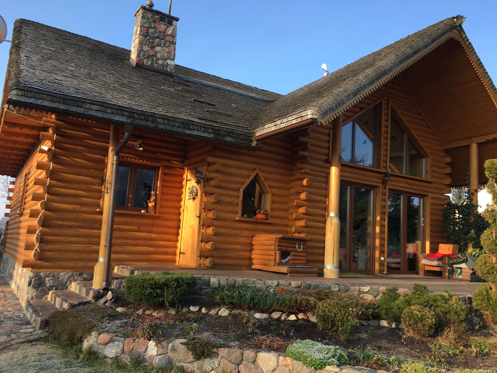 Wykończony dom z bali półokrągłych z dachem krytym gontem drewnianym, widoczny kamienny komin i elementy dekoracyjne dachu, ogródek przed domem.