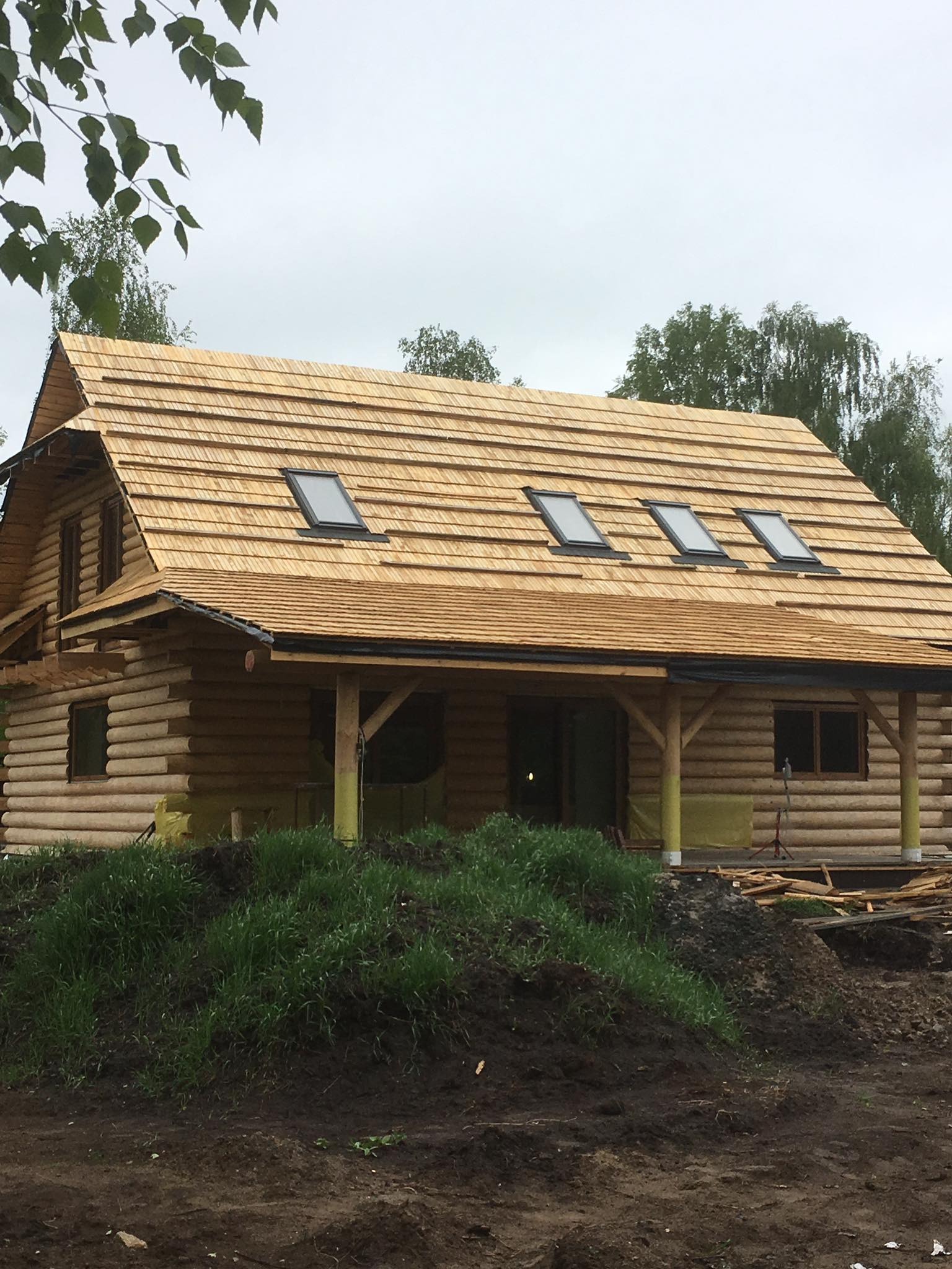 Wznoszony dom z bali w Łomży z drewnianym dachem krytym gontem i czterema oknami dachowymi, widoczne elementy izolacyjne i fragmenty rusztowania.