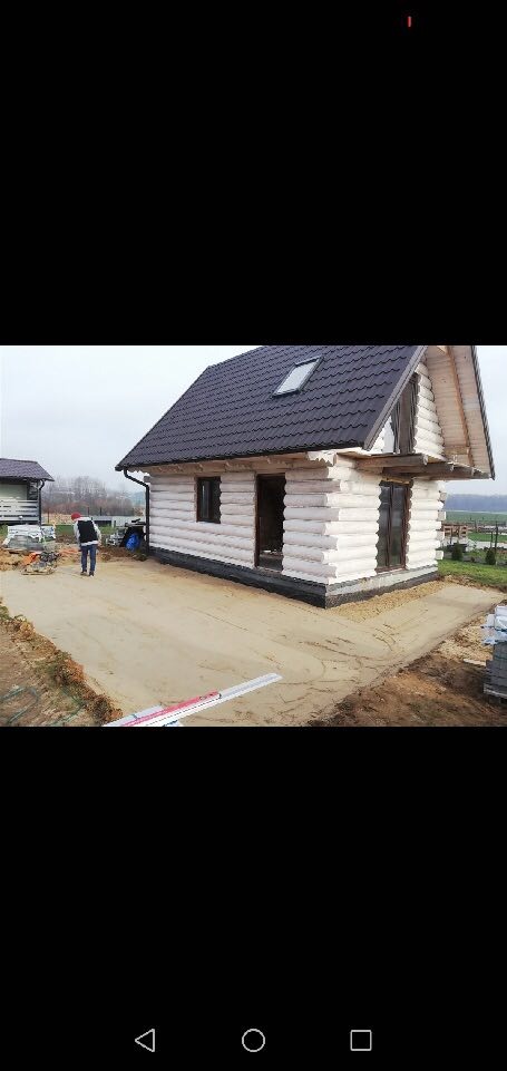 Surowy stan budowy parterowego domu z bali z ciemną dachówką, widoczne przygotowane podłoże i pracownik w oddali.