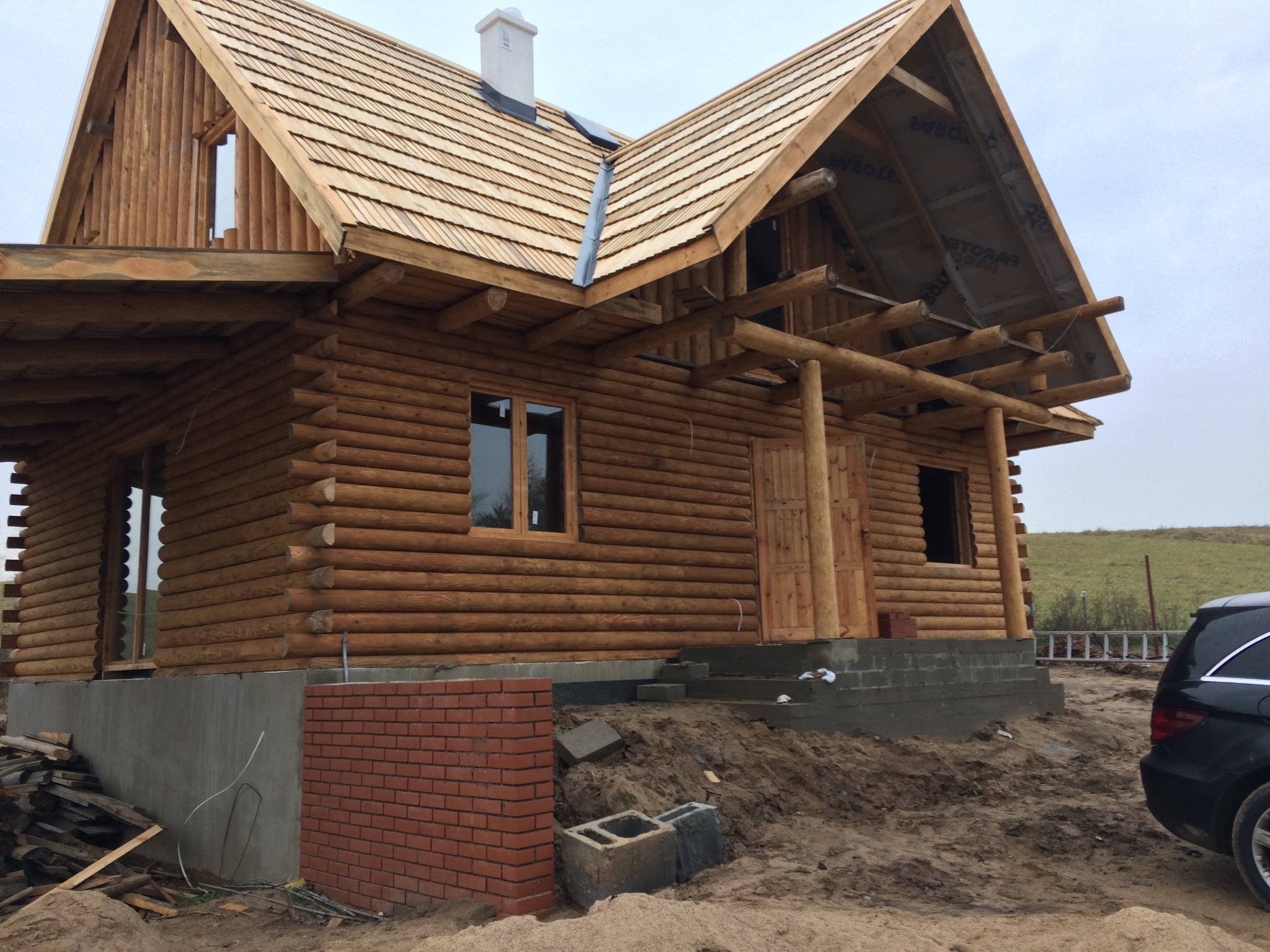 Surowy stan budowy domu z bali z drewnianym gontem na dachu, widoczne elementy konstrukcyjne dachu i niedokończone fundamenty, z boku zaparkowany samochód.