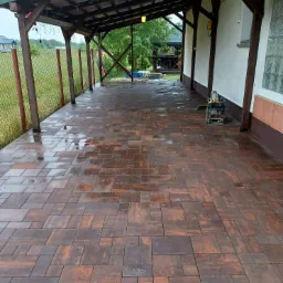 Ułożona, mokra kostka brukowa w odcieniach brązu i pomarańczy pod zadaszeniem drewnianej wiaty, widoczna zagęszczarka w tle.