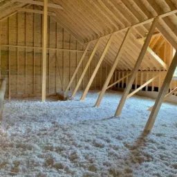 Poddasze w trakcie ocieplania, widoczne drewniane elementy konstrukcyjne i biała warstwa izolacji z wełny mineralnej, częściowo oświetlone światłem dziennym wpadającym przez okno.