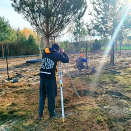 Dwóch pracowników podczas wyznaczania linii ogrodzenia za pomocą niwelatora optycznego; jeden patrzy przez lunetę, drugi wbija słupek w ziemię.