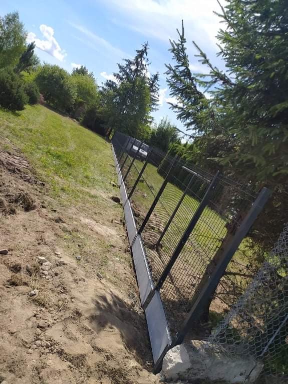 Nowo zainstalowane czarne ogrodzenie panelowe na nierównym terenie, z widoczną ziemią i trawą, częściowo pod kątem, z drzewami w tle.