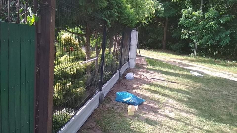 Nowoczesne ogrodzenie panelowe z podmurówką betonową w trakcie montażu, widoczne worki z materiałami budowlanymi na trawie.