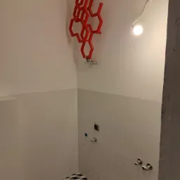 Wykończona łazienka z geometryczną podłogą, białymi płytkami i czerwonym dekoracyjnym grzejnikiem heksagonalnym oraz surowym betonowym filarem po prawej