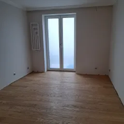 Pusty pokój z nową, jasną drewnianą podłogą, białymi ścianami z listwami przypodłogowymi i sufitowymi, grzejnikiem dekoracyjnym oraz oknem balkonowym.