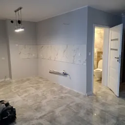 Szara kuchnia w trakcie wykańczania: położone płytki podłogowe imitujące beton, częściowo ułożone płytki ścienne z motywem marmuru, widoczne instalacje hydrauliczne, otwarte drzwi do łazienki...