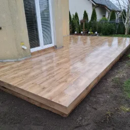 Nowoczesny taras wyłożony płytkami imitującymi drewno, widok z poziomu gruntu, z fragmentem ogrodu i elewacji budynku w tle.