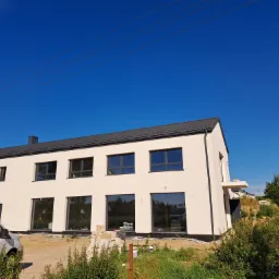 Nowoczesny, dwupiętrowy dom z białym tynkiem zewnętrznym, czarną dachówką i dużymi oknami, widoczny z zewnątrz, z zaparkowanym białym vanem w pobliżu i otoczeniem w trakcie prac wykończeniowych.