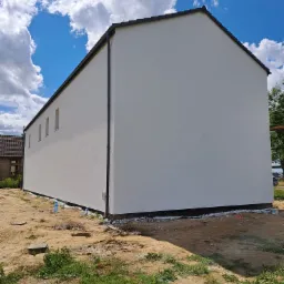 Elewacja domu jednorodzinnego po malowaniu na biało, widoczne okna w pionie, czarne rynny i dachówka, ziemia wokół budynku.