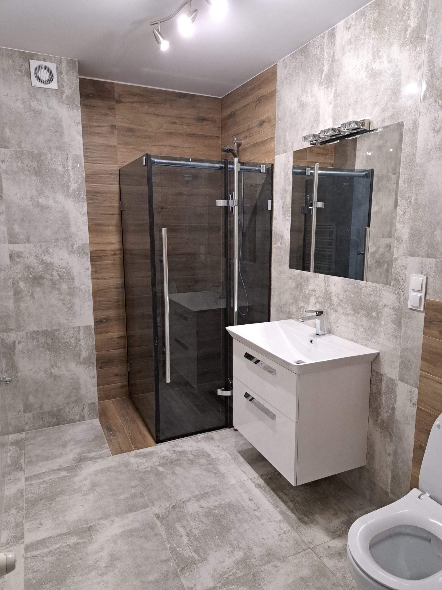 Nowoczesna łazienka z kabiną prysznicową typu walk-in, umywalką z szafką i toaletą; szaro-brązowe płytki imitujące beton i drewno, oświetlenie punktowe i kryształowe nad lustrem.