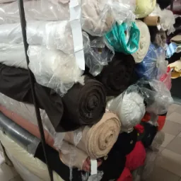 Stos różnokolorowych bel materiałów w plastikowych osłonach, składowanych w magazynie, widoczne odcienie bieli, beżu, brązu, czerni, turkusu i żółci.