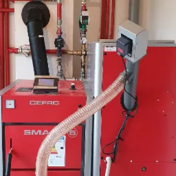 Czerwony piec CO marki Defro Smart z podajnikiem, widoczne rury instalacji grzewczej i komin, elementy sterujące i wyświetlacz.