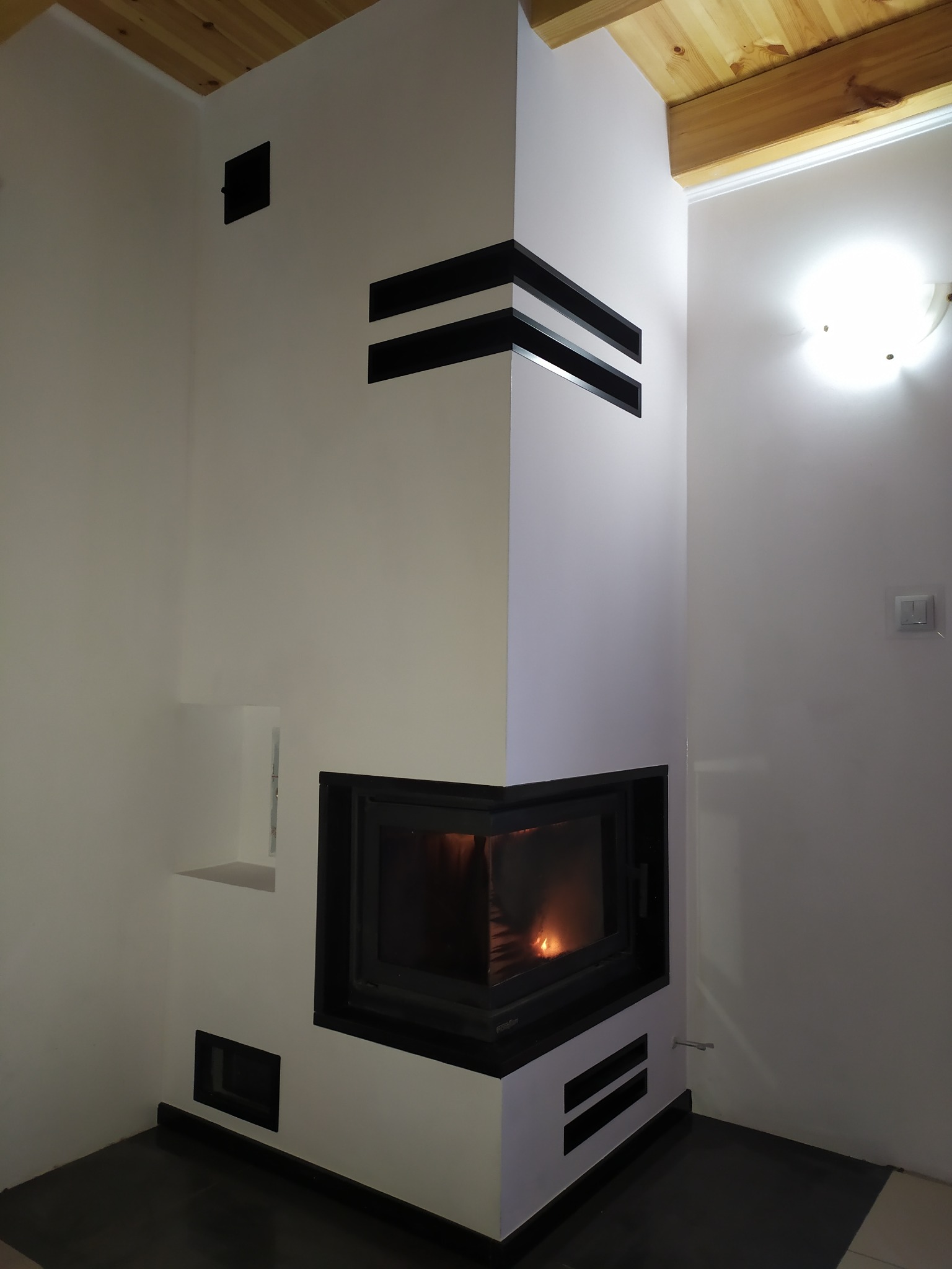 Biały narożny z wkładem kominkowym, obudowany minimalistyczną formą z czarnymi, poziomymi akcentami, widoczny płomień w palenisku i fragment drewnianego sufitu.