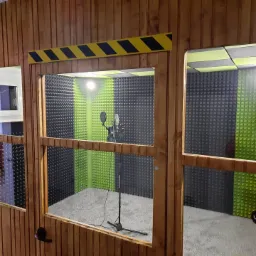 Wnętrze kabiny akustycznej z mikrofonem na statywie, wykończonej drewnem i panelami akustycznymi w kolorach szarym i zielonym, widok przez okno z żółto-czarnym pasem ostrzegawczym na górze.