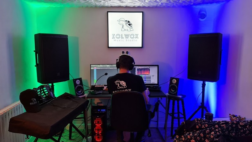 Osoba w słuchawkach siedzi przed dwoma monitorami z oprogramowaniem do edycji audio, w tle głośniki, logo studia muzycznego z żółwiem i oświetlenie LED w kolorach zielonym i niebieskim.