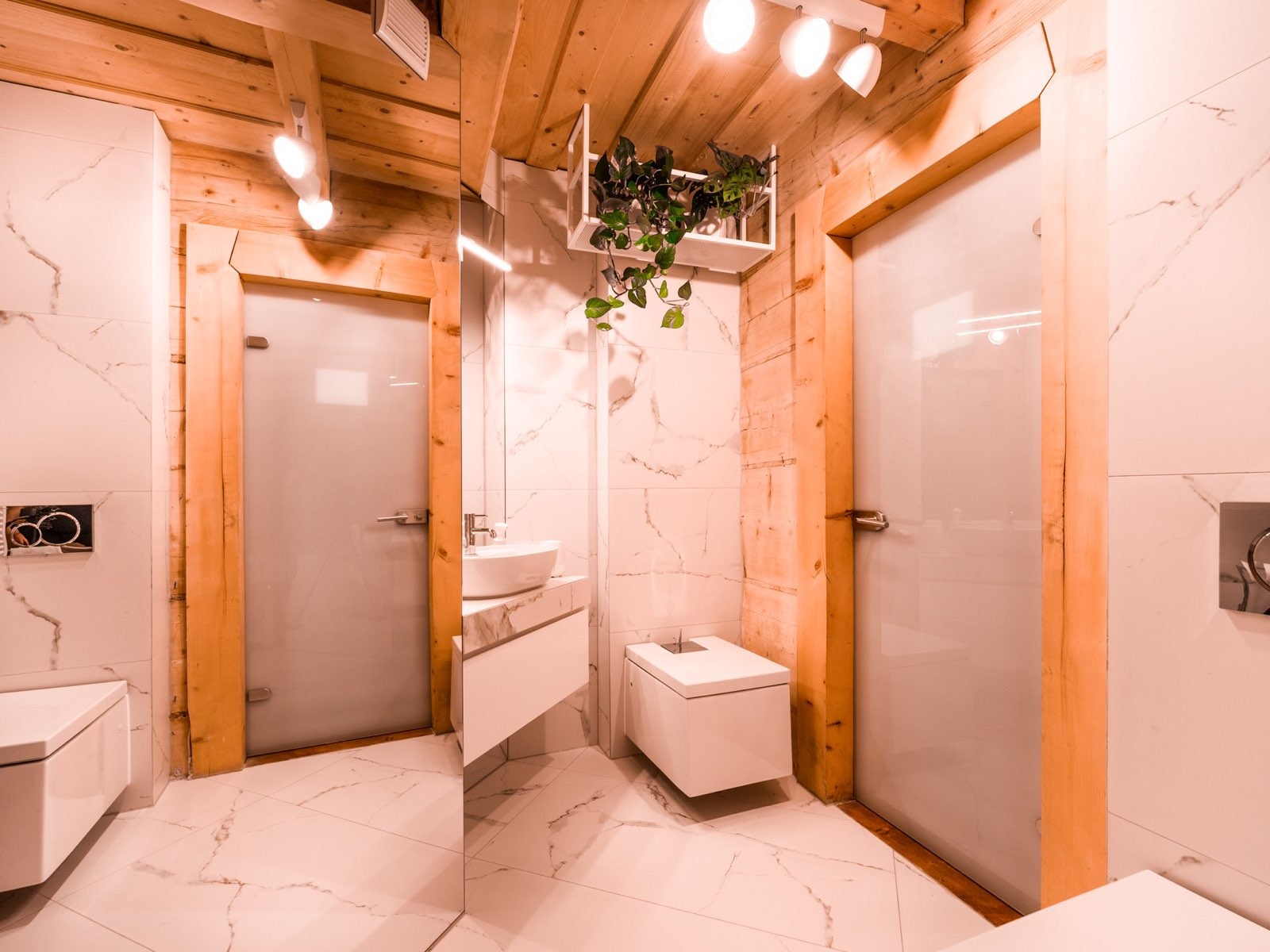 Nowoczesna łazienka z białymi płytkami imitującymi marmur, drewnianymi elementami wykończeniowymi, szklanymi drzwiami i roślinami w dekoracyjnej półce.