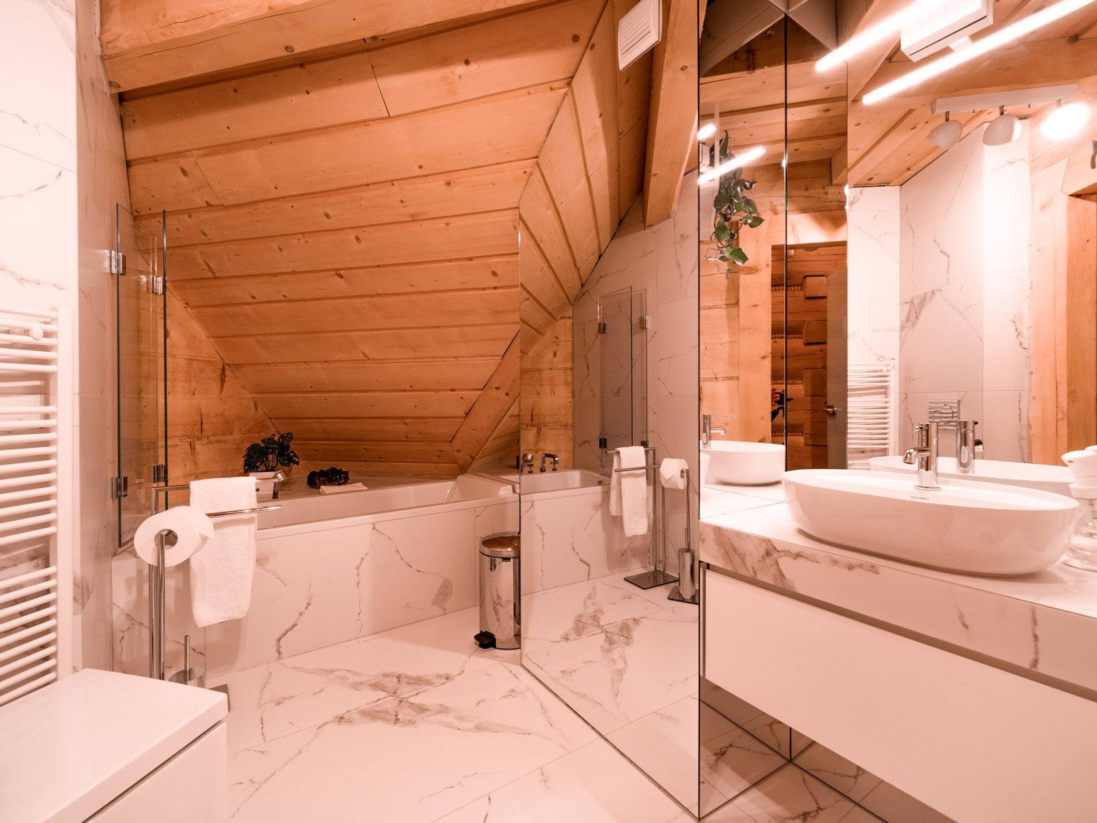 Jasna łazienka na poddaszu z drewnianym skośnym sufitem, białymi marmurowymi płytkami, owalną umywalką i lustrem na całej ścianie.