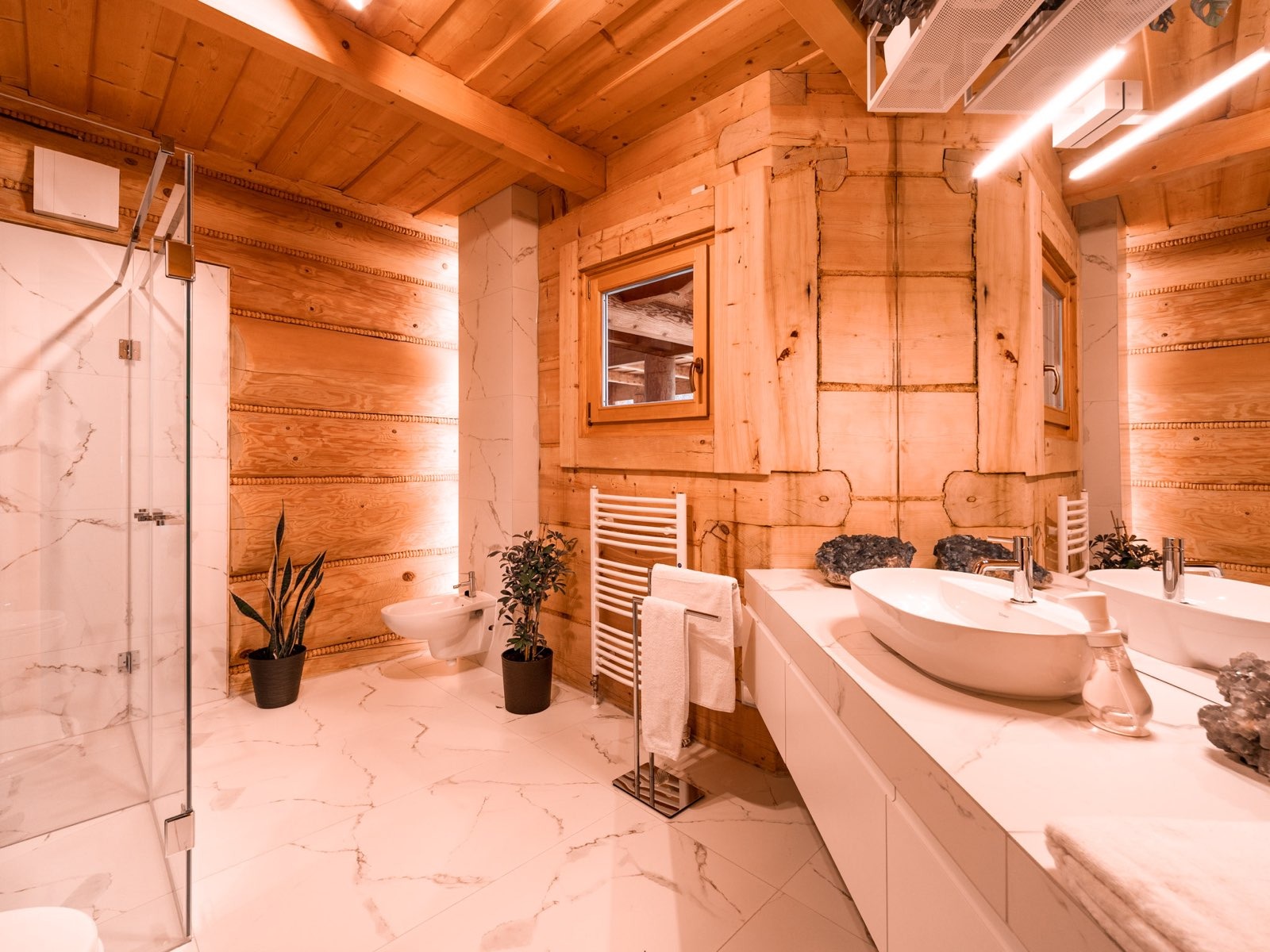 Nowoczesna łazienka w stylu góralskim z drewnianymi ścianami, marmurową podłogą i białymi sanitariatami, akcentowana roślinami doniczkowymi i oświetleniem LED.