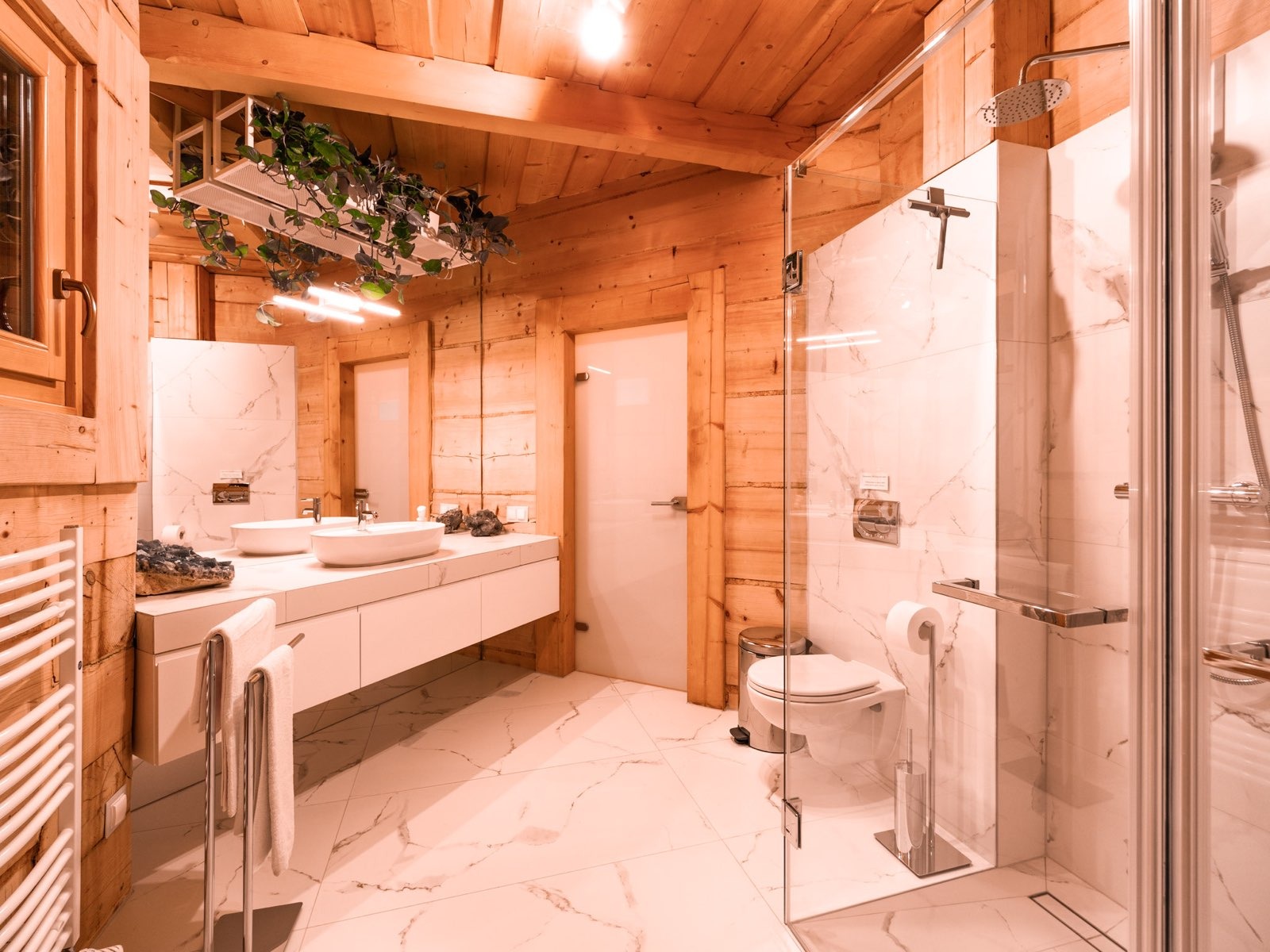 Jasne, drewniane wnętrze łazienki z marmurową podłogą i kabiną prysznicową ze szkła, podwójna umywalka z dekoracyjnymi kamieniami i wiszącymi roślinami pod sufitem.