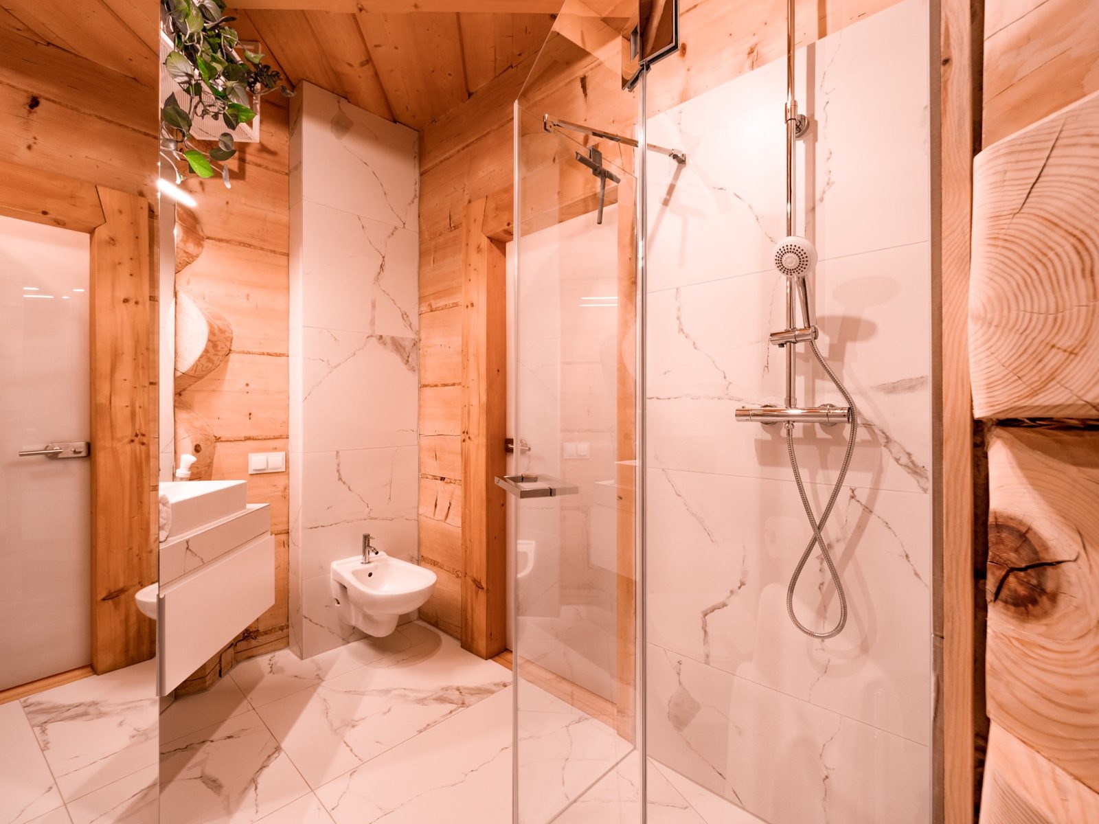 Nowoczesna łazienka w stylu rustykalnym: drewniane ściany i sufit, białe marmurowe płytki, szklana kabina prysznicowa, biała umywalka i bidet.