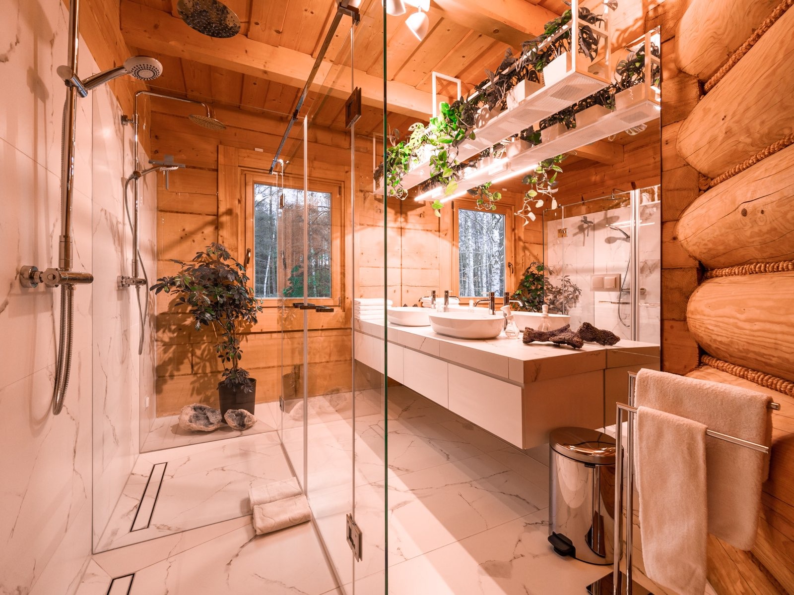 Drewniana łazienka z kabiną prysznicową ze szkła, białymi umywalkami i roślinami doniczkowymi, widok z perspektywy wejścia.
