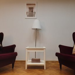 Gabinety Psychoterapii ARIADNA Kraków Długa - Pokój terapeutyczny z dwoma bordowymi fotelami typu uszak, białym stolikiem z lampką i obrazkiem okna na ścianie.
