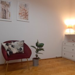 Gabinety Psychoterapii ARIADNA Kraków Długa - Pokój z bordowym fotelem, geometrycznymi poduszkami, rośliną w szarej doniczce, komodą z trzema szufladami i lampką nocną, udekorowany dwoma obrazkami na ścianie.