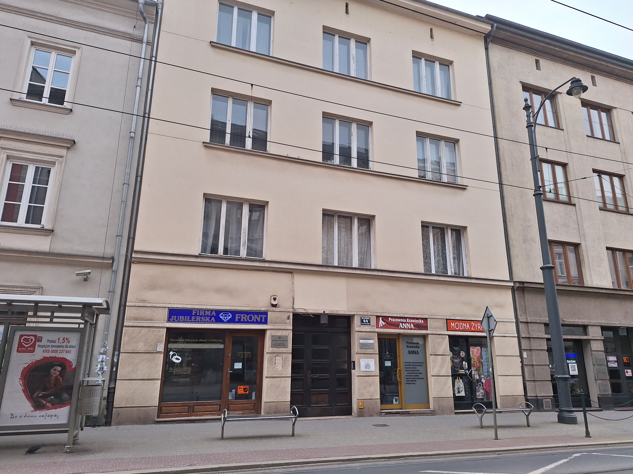 Fasada kamienicy z kilkoma lokalami usługowymi na parterze, w tym pracownia krawiecka z szyldem 'Pracownia Krawiecka ANNA' i informacjami o świadczonych usługach.