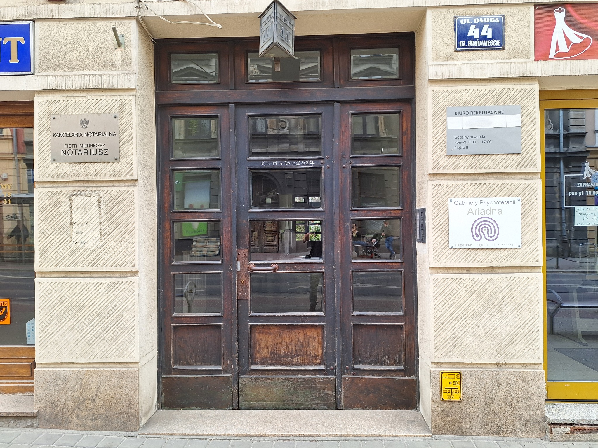 Wejście do budynku z drewnianymi drzwiami z szybami, tabliczką 'Gabinety Psychoterapii Ariadna' oraz adresem 'Ul. Długa 44/6', widoczny napis kredą na drzwiach 'K+M+B=2024'.