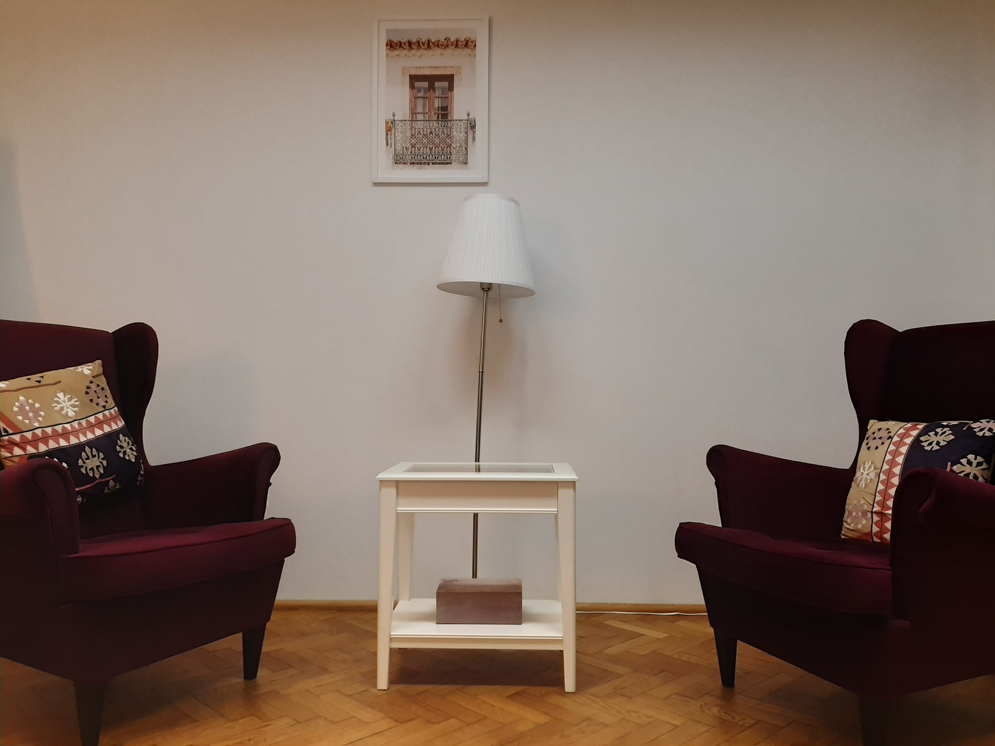 Pokój terapeutyczny z dwoma bordowymi fotelami typu uszak, białym stolikiem z lampką i obrazkiem okna na ścianie.