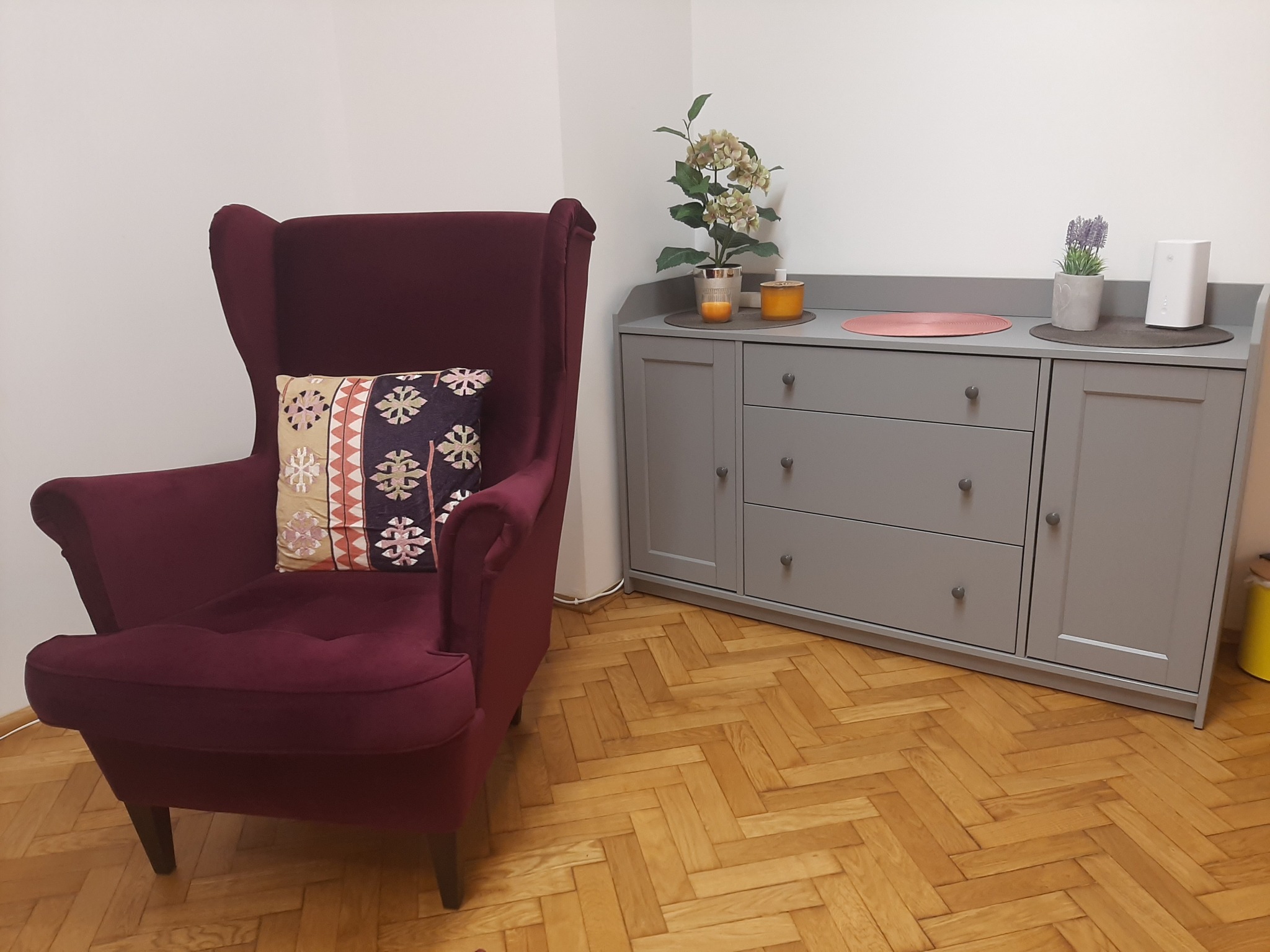 Burgundowy fotel typu uszak z wzorzystą poduszką stoi obok szarej komody z dekoracjami roślinnymi i świecami na blacie, na parkiecie ułożonym w jodełkę.