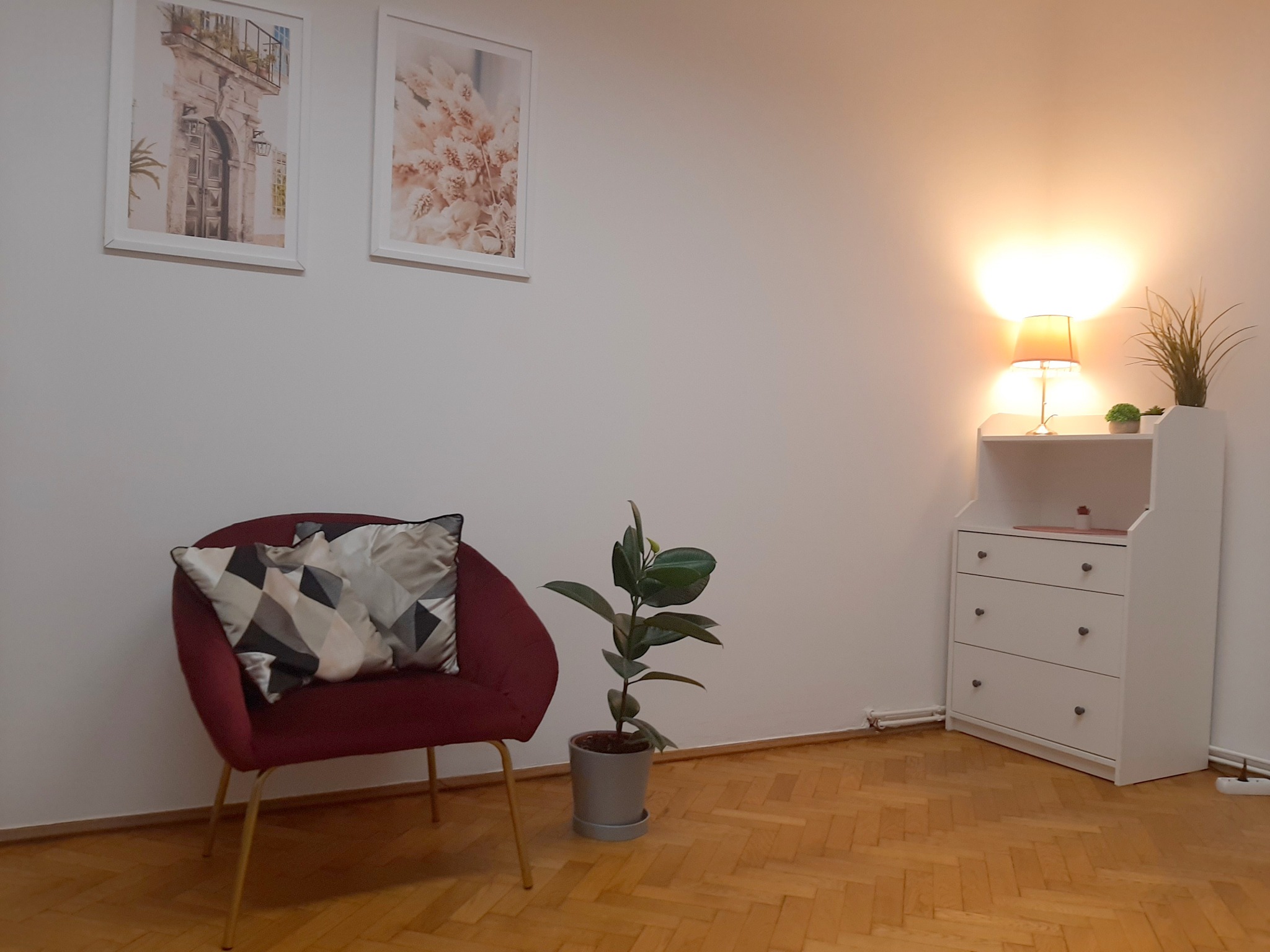 Pokój z bordowym fotelem, geometrycznymi poduszkami, rośliną w szarej doniczce, komodą z trzema szufladami i lampką nocną, udekorowany dwoma obrazkami na ścianie.