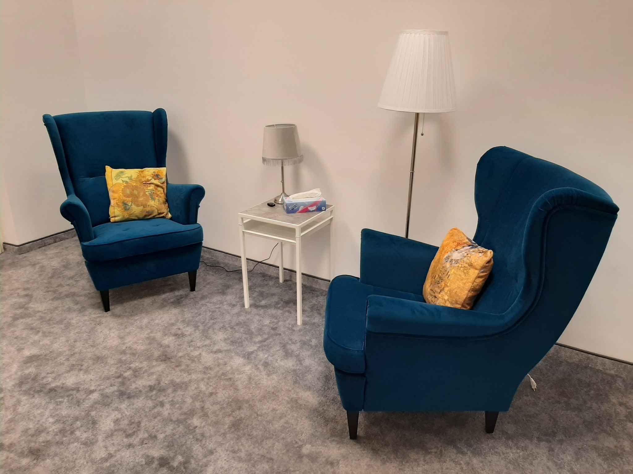 Pokój z dwoma turkusowymi fotelami typu uszak, żółtymi poduszkami z motywem kwiatowym, stolikiem z chusteczkami i dwiema lampami o różnej wysokości, na tle jasnych ścian i szarej wykładziny.