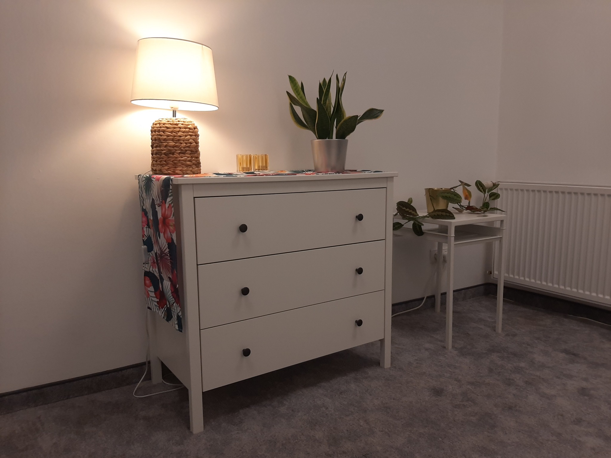 Jasna komoda z trzema szufladami, na której stoi lampka z abażurem z rafii, roślina doniczkowa w srebrnej osłonce, dwa małe szklane wazoniki oraz serwetka w kwiaty. W tle stolik z rośliną i grzejnik.